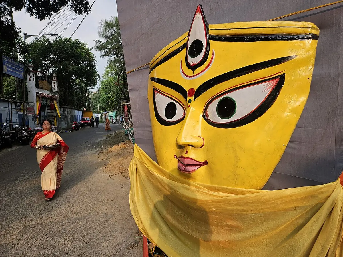 A woman in a red-bordered white sari walks past a giant Durga face in Kolkata.